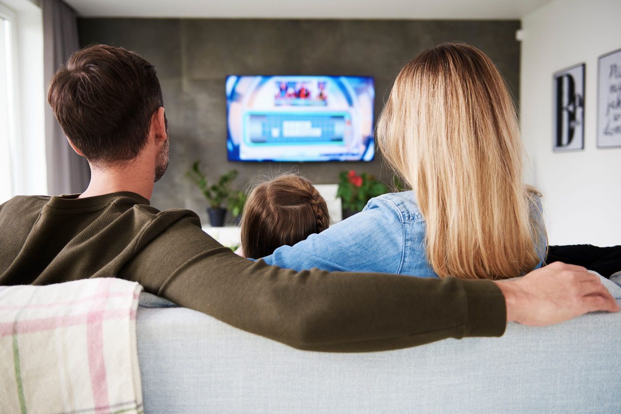 A rear view of a family of three - mum, daughter and mother - sat on a sofa and watching the television.