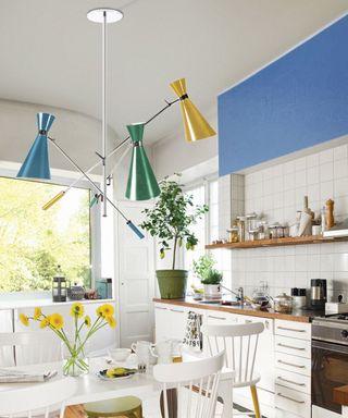 A bright, airy kitchen with a Scandinavian-inspired design. The walls and cabinetry are crisp white, with a backsplash of square white tiles. Above the table hangs a striking mid-century modern chandelier with three cone-shaped lampshades in green, yellow, and blue, attached to black metal rods extending from a chrome ceiling mount