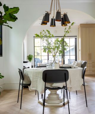 Round dining table with white cloth, dark chairs, and abundance of foliage in bright space with modern spot ceiling lights