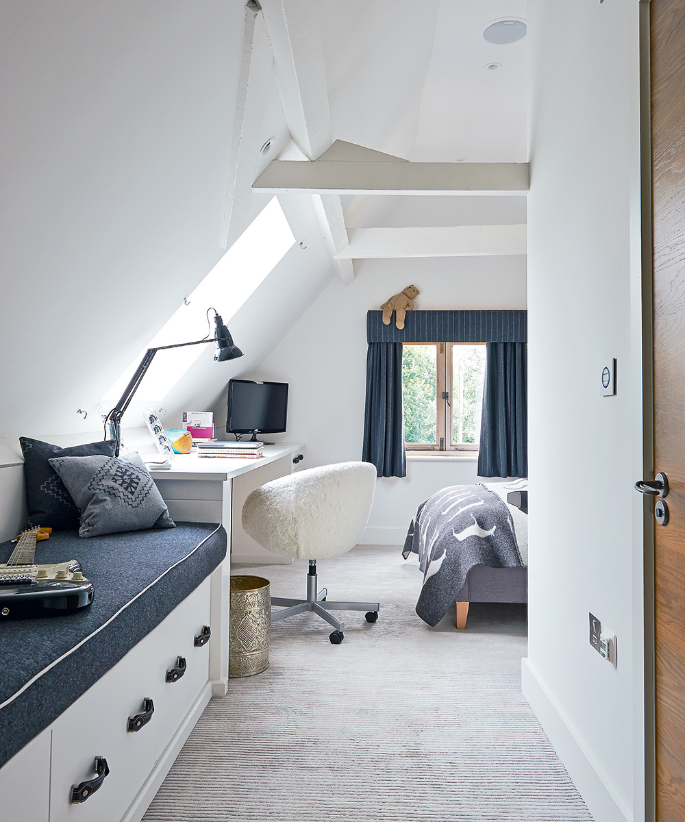 a white attic bedroom with pale gray carpet, white wooden furniture and navy blue cushions, bedding and drapes illustrating coordinated small bedroom ideas for kids.