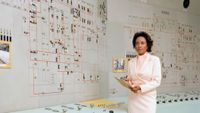 A Black woman wearing a pale pink skirt suit set stands in front of a control board with diagrams.