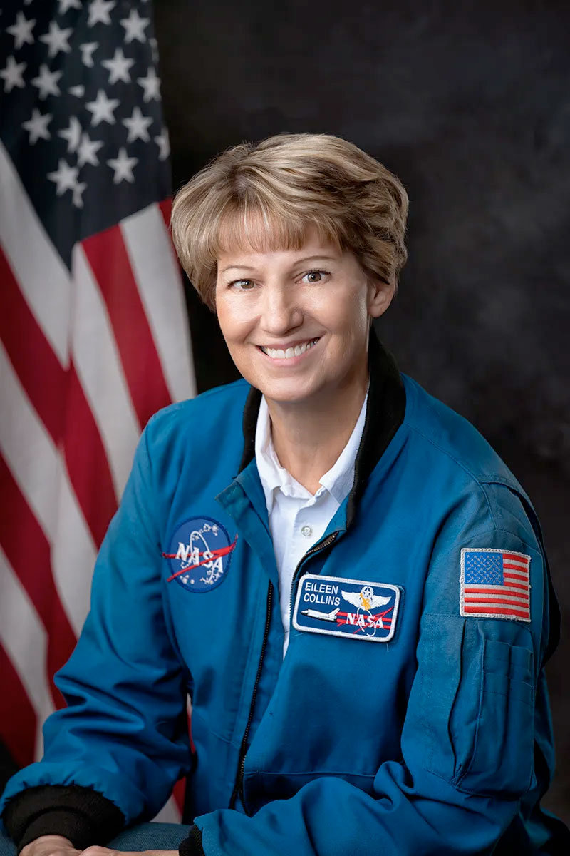 a smiling woman in a blue flight suit sits in front of an American flag