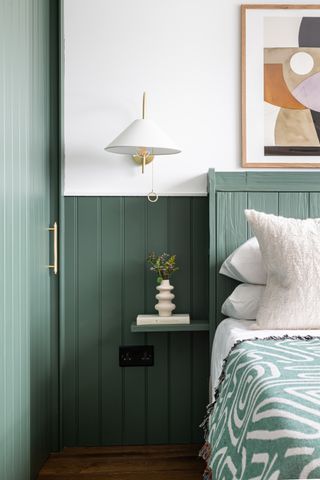 green bedroom with painted tongue & groove, white walls, white wall light, artwork, small shelf, wooden floors, green abstract blanket
