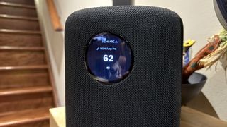 The WiiM Sound, in a home, on a varnished wooden table, showing the illuminated front circular screen. It currently displays the volume, and information that its music source is the HDMI port of a WiiM Amp Pro.