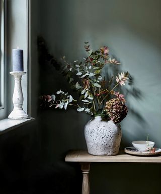 Close up of seasonal flowers and branches in cream ceramic vase on shelf, blue painted walls windowsill with candle in holder