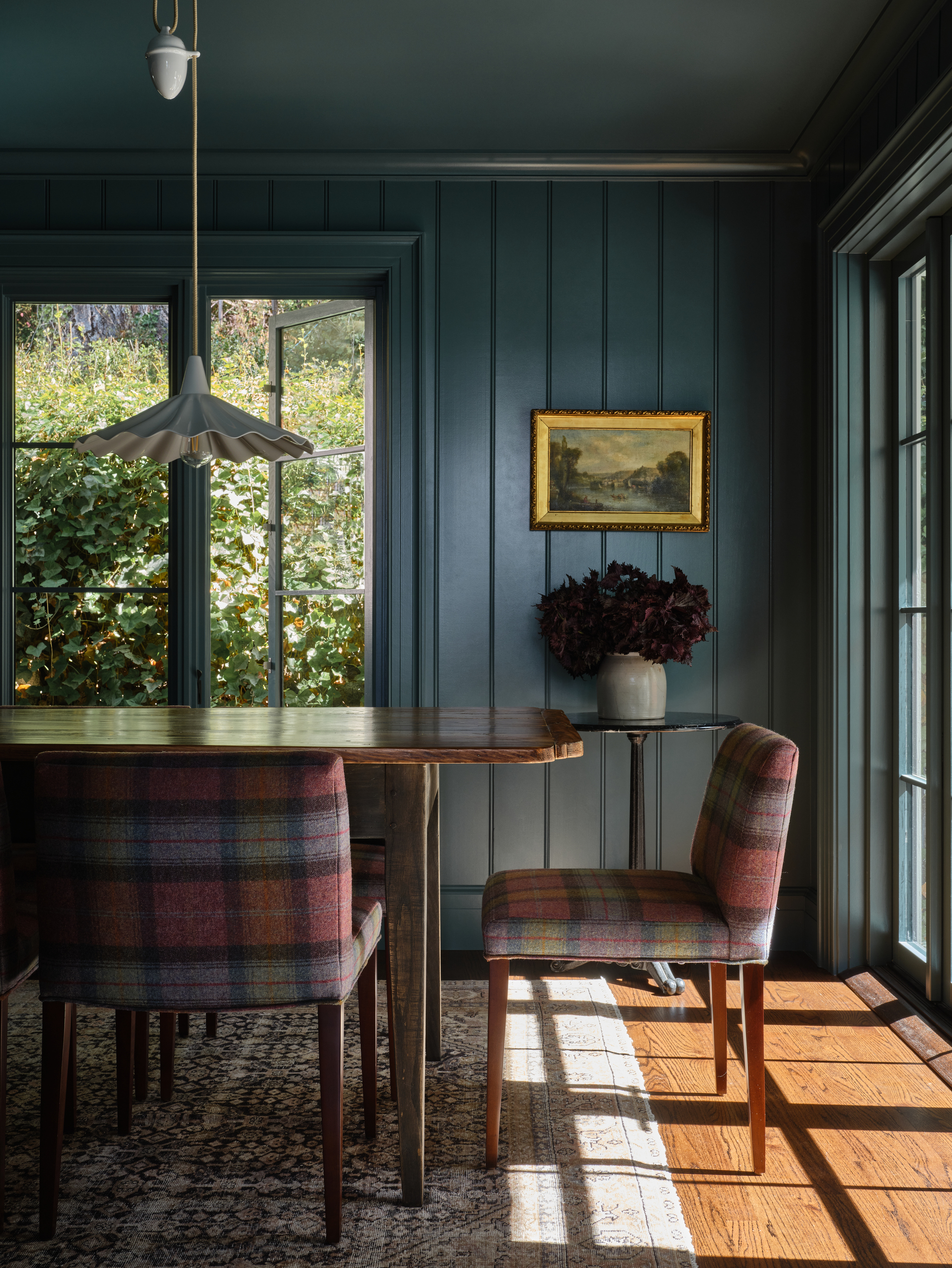 A dark teal and navy blue, panel-drenched dining room with hardwood floors and a wooden dining table. There are pink plaid dining chairs around the table.