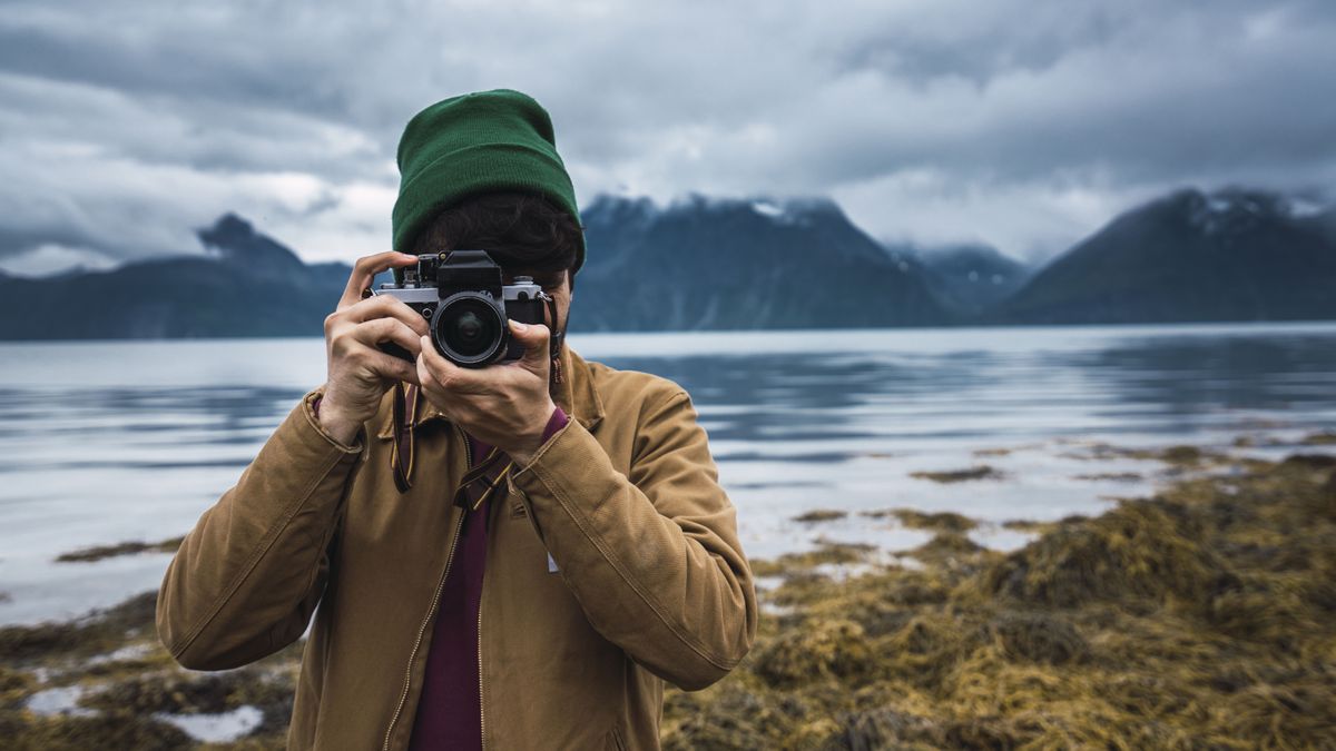 Best beginner cameras: Image shows person in woolly hat taking picture with water and mountains behind them