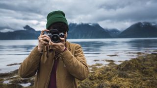 Best beginner cameras: Image shows person in woolly hat taking picture with water and mountains behind them