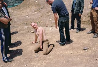 A shirtless man kneels on dry, dusty ground looking upward at a group of men standing around him outdoors.