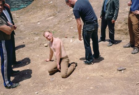 A shirtless man kneels on dry, dusty ground looking upward at a group of men standing around him outdoors.