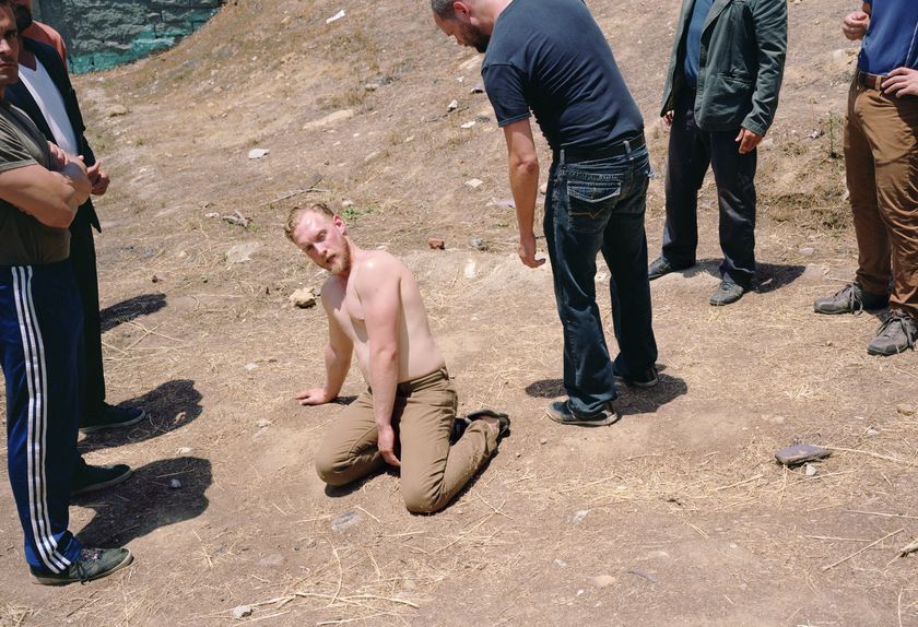 A shirtless man kneels on dry, dusty ground looking upward at a group of men standing around him outdoors.