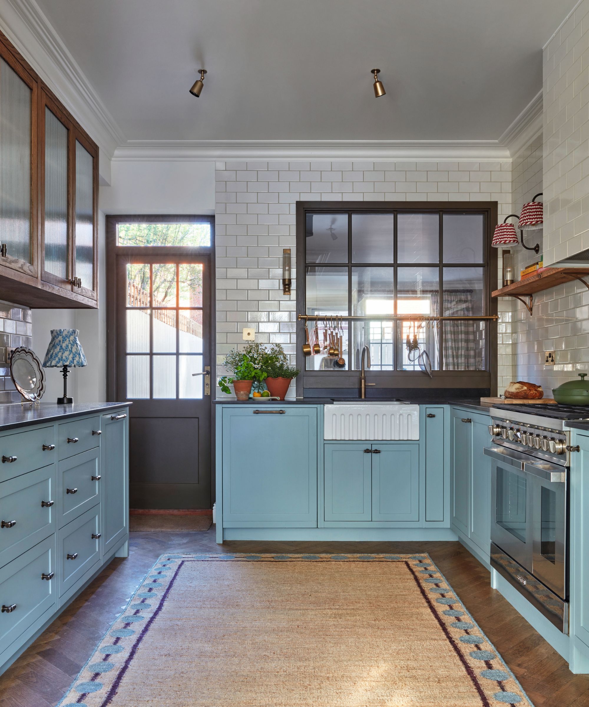 A kitchen with blue cabinets, white subway tiles covering the walls, a large jute rug on the floor, and glass-fronted cabinets on the upper wall