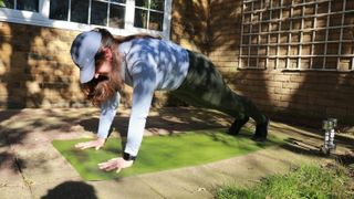 Writer Marina Leiva García performs a plank outside on a mat. She is supporting herself on her hands and tiptoes, with the rest of her body elevated off the ground. It is sunny and the long shadows of trees fall over her body.