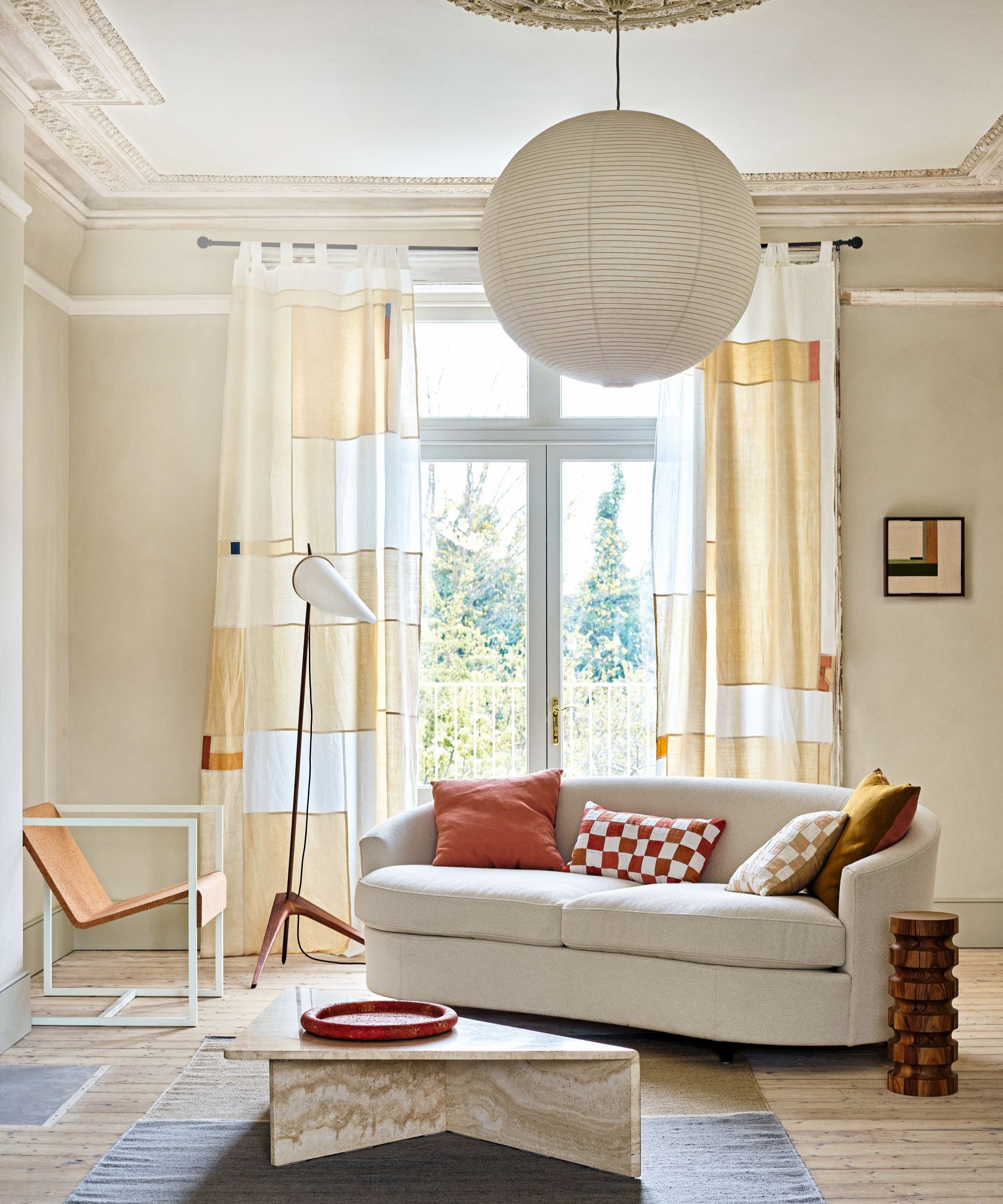 a living room with cream painted walls, long patchwork style drapes in front of the windows, a white couch with colorful pillows, an accent chair, a floor lamp, a coffee table, and a paper ceiling pendant
