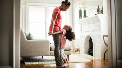 A small child stands on top of the feet of her smiling mom and looks up at her in their living room.