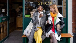 Two women dressed in bright colors sitting on bench outside coffee shop with takeaway coffee cups in hand