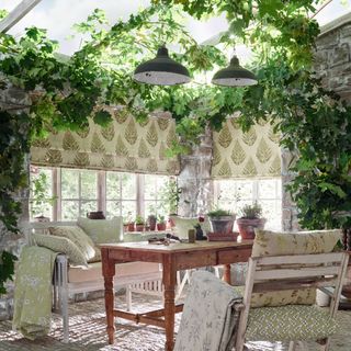conservatory/garden room with fabric blinds, foliage woven into the beams, bench, rustic table and chairs with cushions and throws, plant pots, pair of matching green enamel pendants