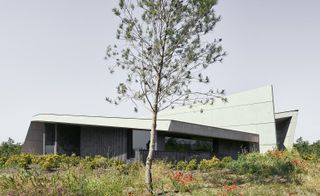 Exterior view of Crematorio Igualada featuring grey and white colours photographed behind bushes and a mini tree