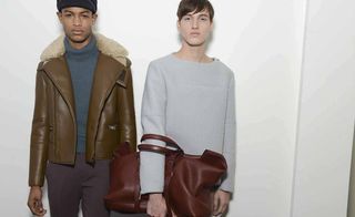 Two male models wearing looks from Gucci's collection. One model is wearing a black hat, dark coloured trousers, green roll neck jumper and brown jacket with fur. The other model is wearing a light grey sweater and is holding a large dark red bag