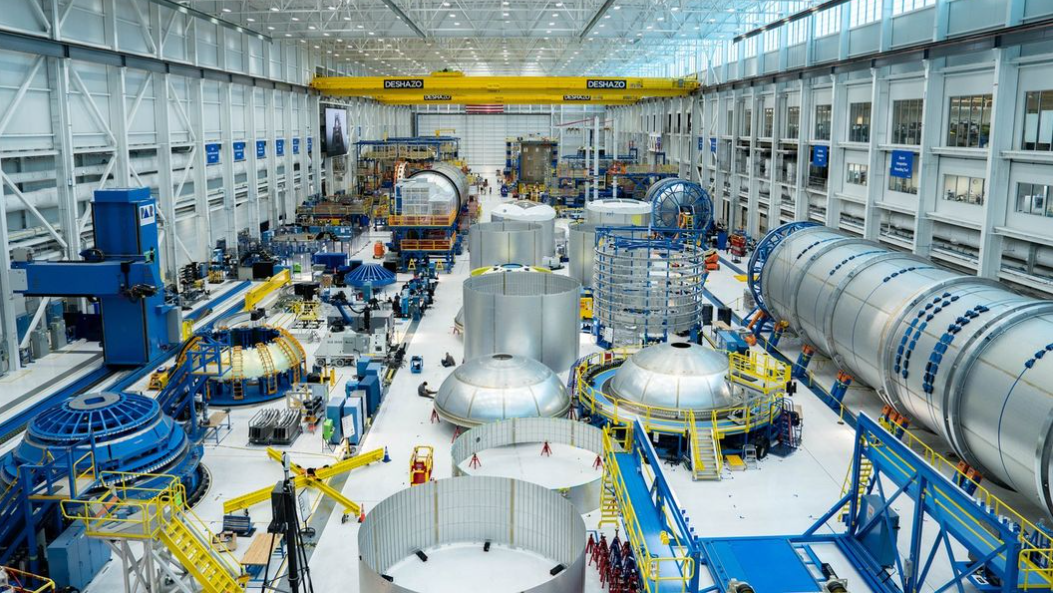 Cylindrical and dome pieces of metal are organized and laid out on the floor of a large factory. A long fuselage lays on the right.