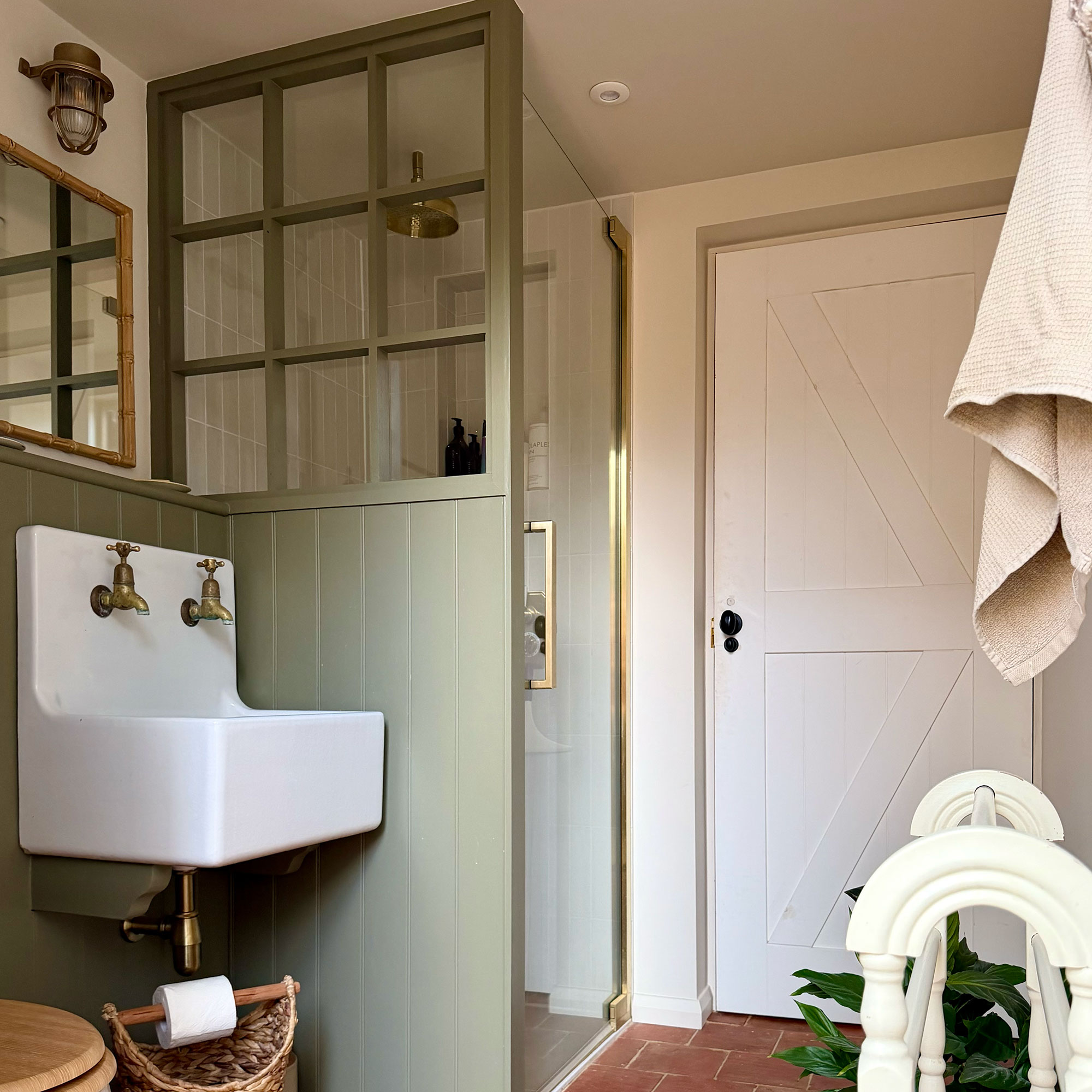 Cream bathroom with green wall panelling on wall and shower enclosure with glass front and gold hardware beside country sink