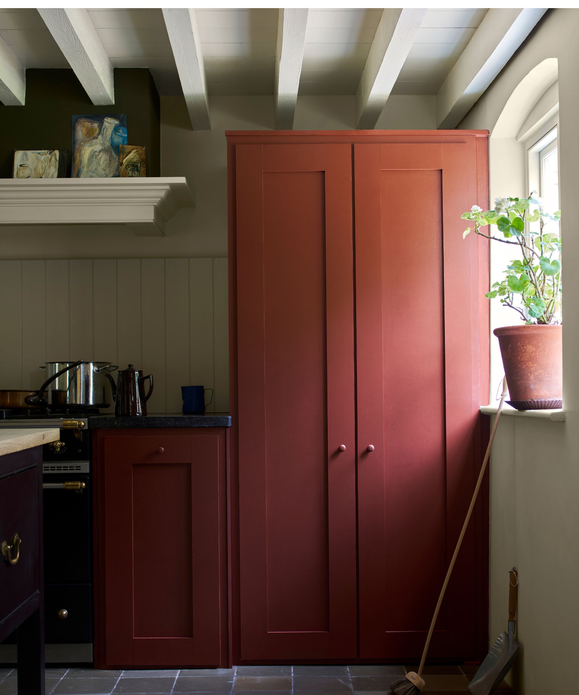 Kitchen with beamed ceiling, Harrisay by Farrow &amp;amp; Ball painted cupboard and cabinetry, a tiled slate floor, an arched window with a potted plant on the sill.
