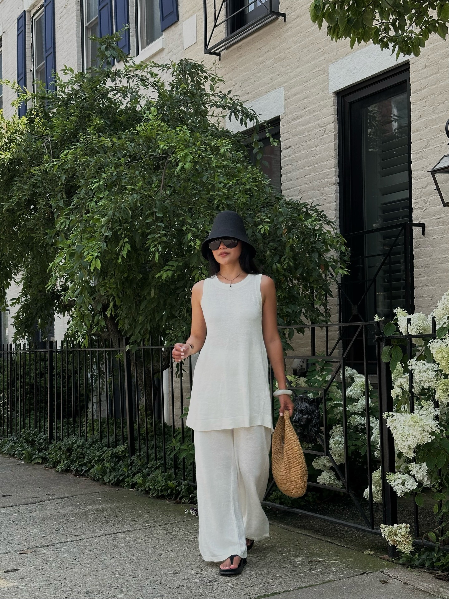 Woman wears raffia hat, white top and linen trousers, carries woven bag