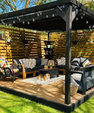 Black decked pergola design with beagle sat under shade and rattan garden furniture