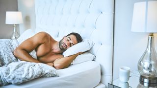 A bare chested man sleeps on his side with his hand propped under one pillow