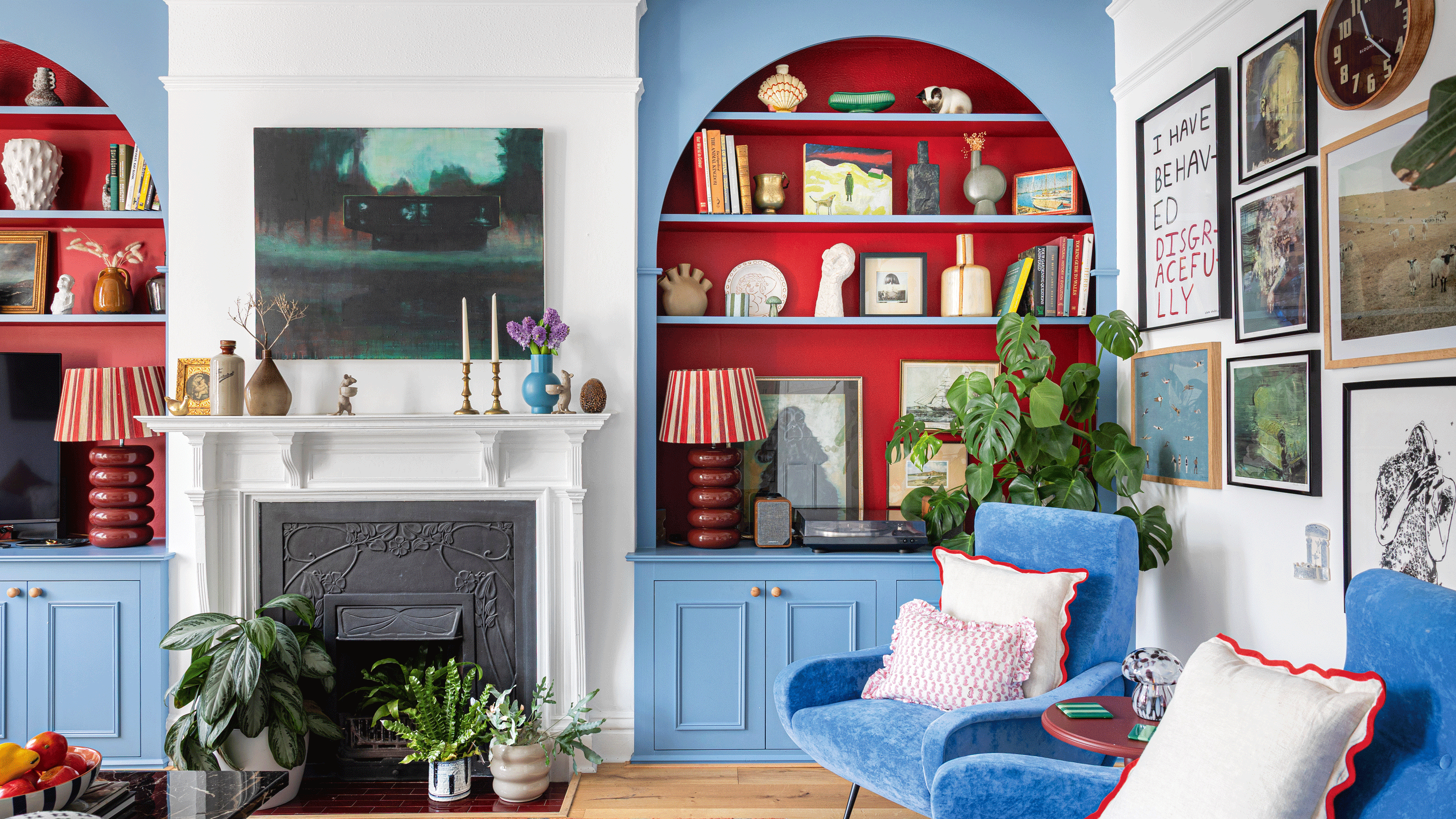 a living room with bright blue painted alcove shelving with bold contrasting red recessed shelves with a gallery wall, fireplace and plenty of colour