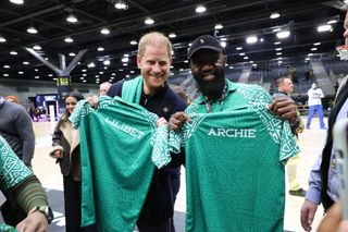 Prince Harry and a man holding green shirts that say Archie and Lilibet at a basketball arena