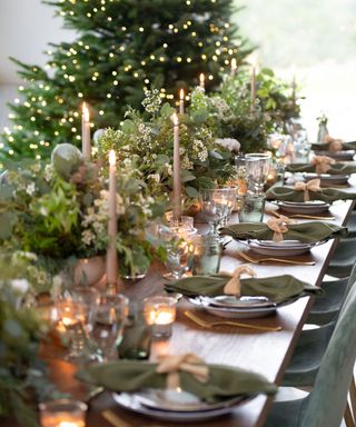 Large christmas dining table decorated with green foliage, candles, tableware, christmas tree in background