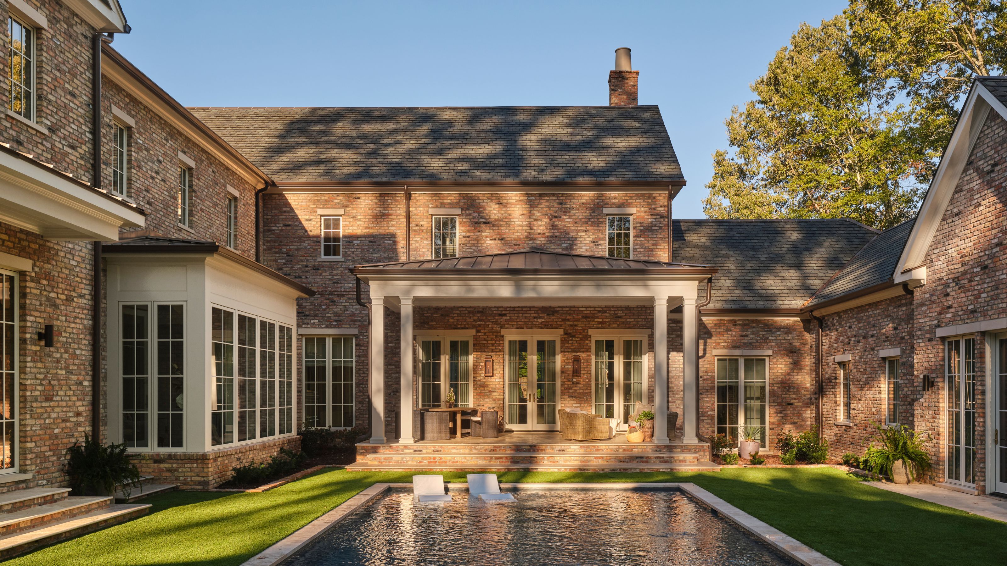 a large brick Georgian style new build with a pool, porch, and soft neutral french doors and windows all facing into the pool