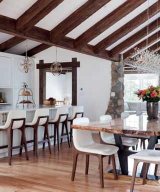 A kitchen with white walls and ceiling, dark wooden beams on the ceiling, white kitchen island and cabinets, and a wooden dining table with white seats underneath