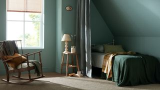 A tranquil bedroom painted in muted teal tones. Sunlight streams through a tall window with a woven blind, casting soft shadows across a natural fibre carpet. slim curtain partially screens a small bed tucked under a sloped ceiling, dressed in green bedding and a mustard throw. A simple wooden stool holds a lamp and ceramic jug beside the bed.