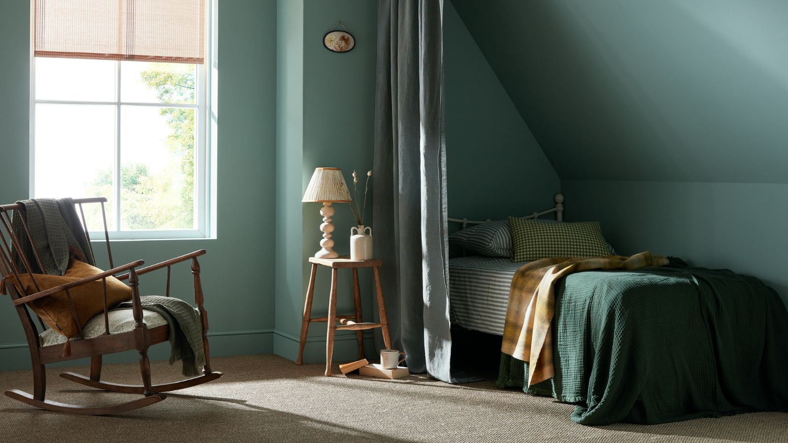 A tranquil bedroom painted in muted teal tones. Sunlight streams through a tall window with a woven blind, casting soft shadows across a natural fibre carpet. slim curtain partially screens a small bed tucked under a sloped ceiling, dressed in green bedding and a mustard throw. A simple wooden stool holds a lamp and ceramic jug beside the bed.