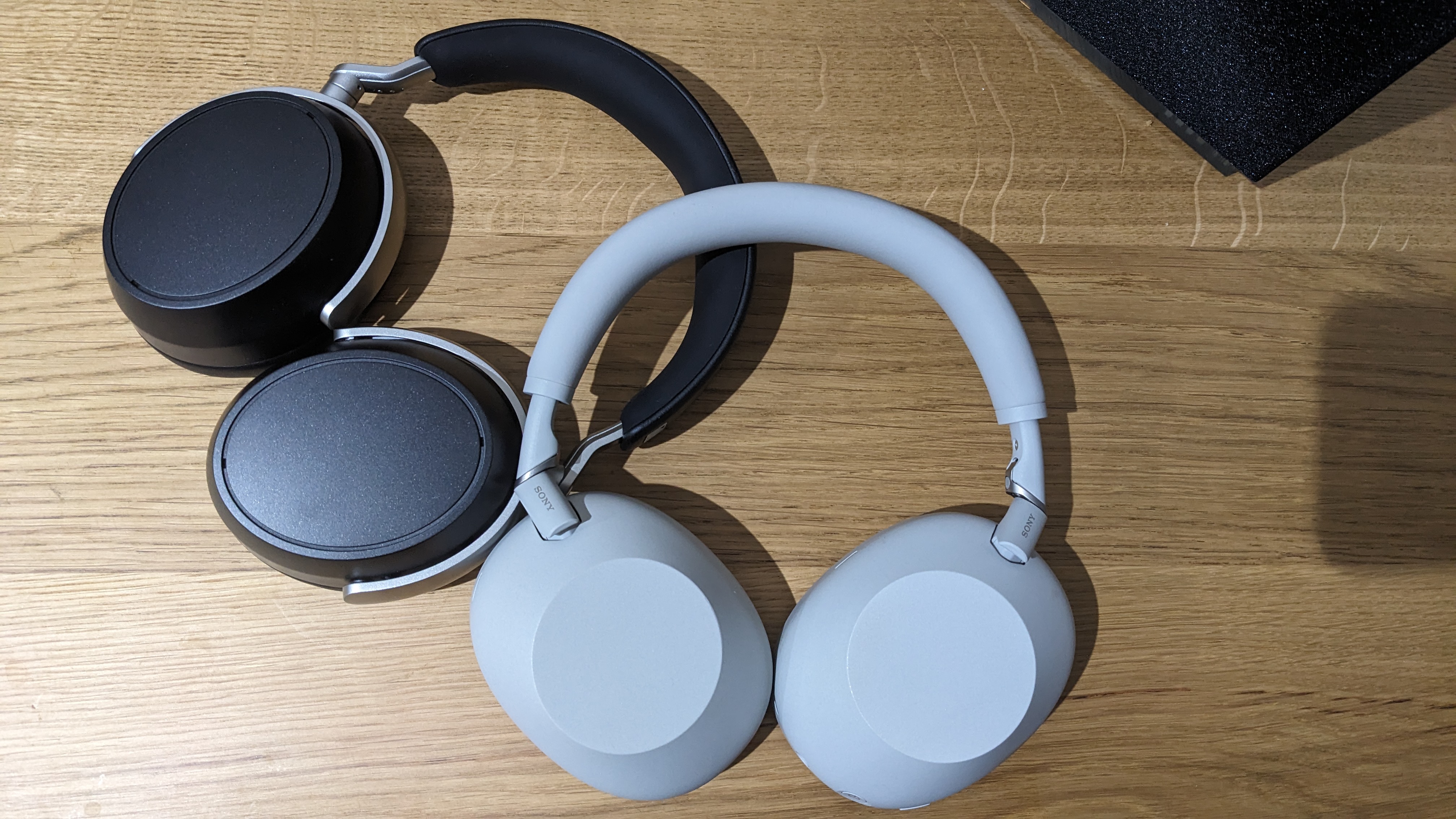 A black pair of Sennheiser HDB 630 and white pair of Sony WH-1000XM6 over-ear headphones lying on a wooden surface, shot from above.