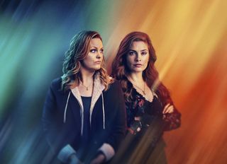 The For Her Sins key art, showing Laura (Jo Joyner) and Emily (Rachel Shenton) standing side by side in front of a multicoloured gradient background of rainbow-style colours. Laura is looking over her shoulder at Emily, while Emily is looking straight into the camera.