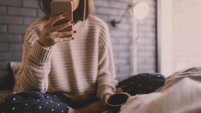 Photo of smiling young woman drinking first morning coffee in her bed, and reading online news on a smart phone