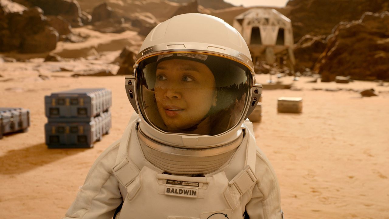 Close up of a female astronaut on Mars. In the background there are several containers and a white building.