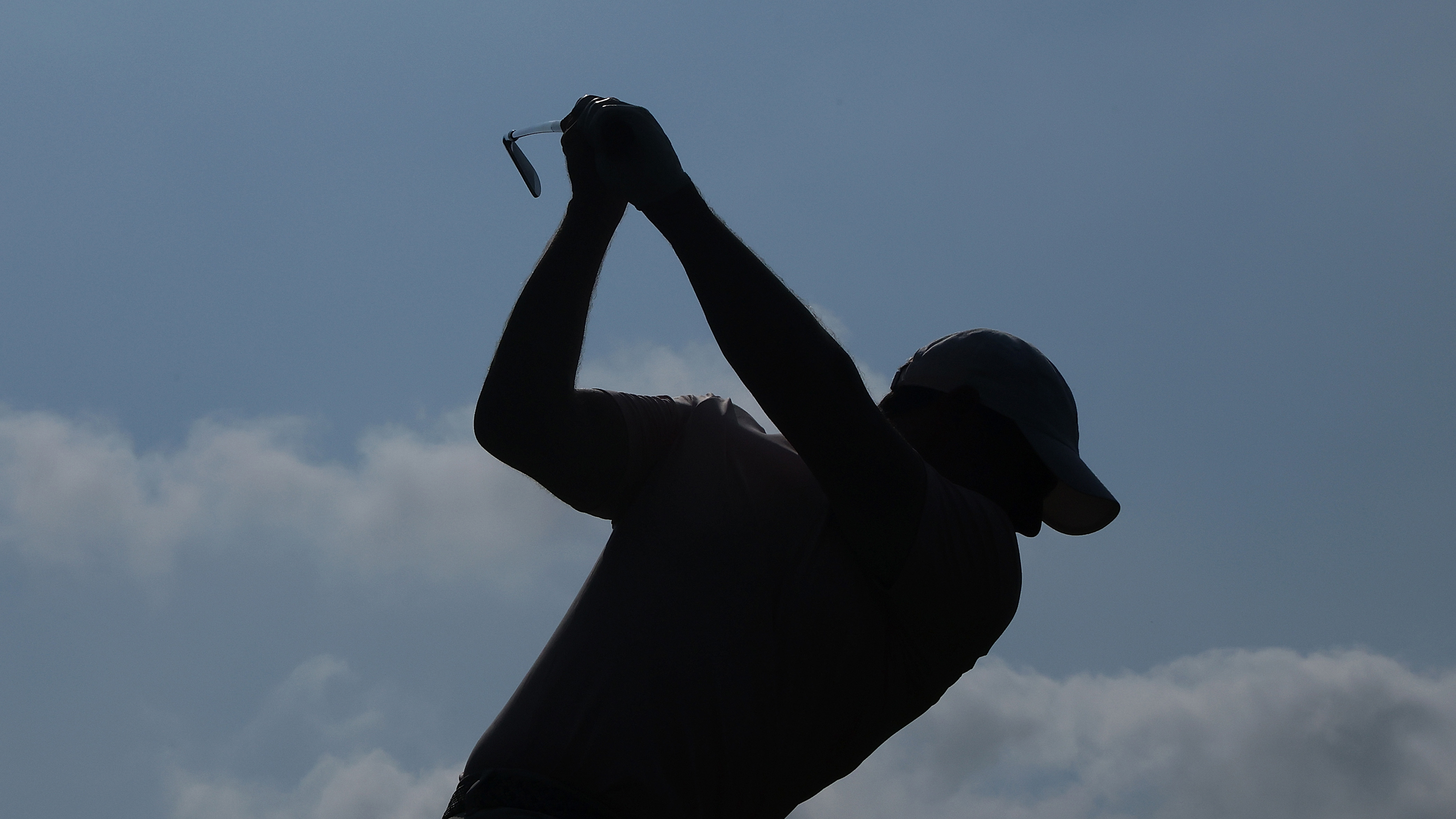 Silhouette of Rory McIlroy at the top of his backswing with an iron, against a blue sky background