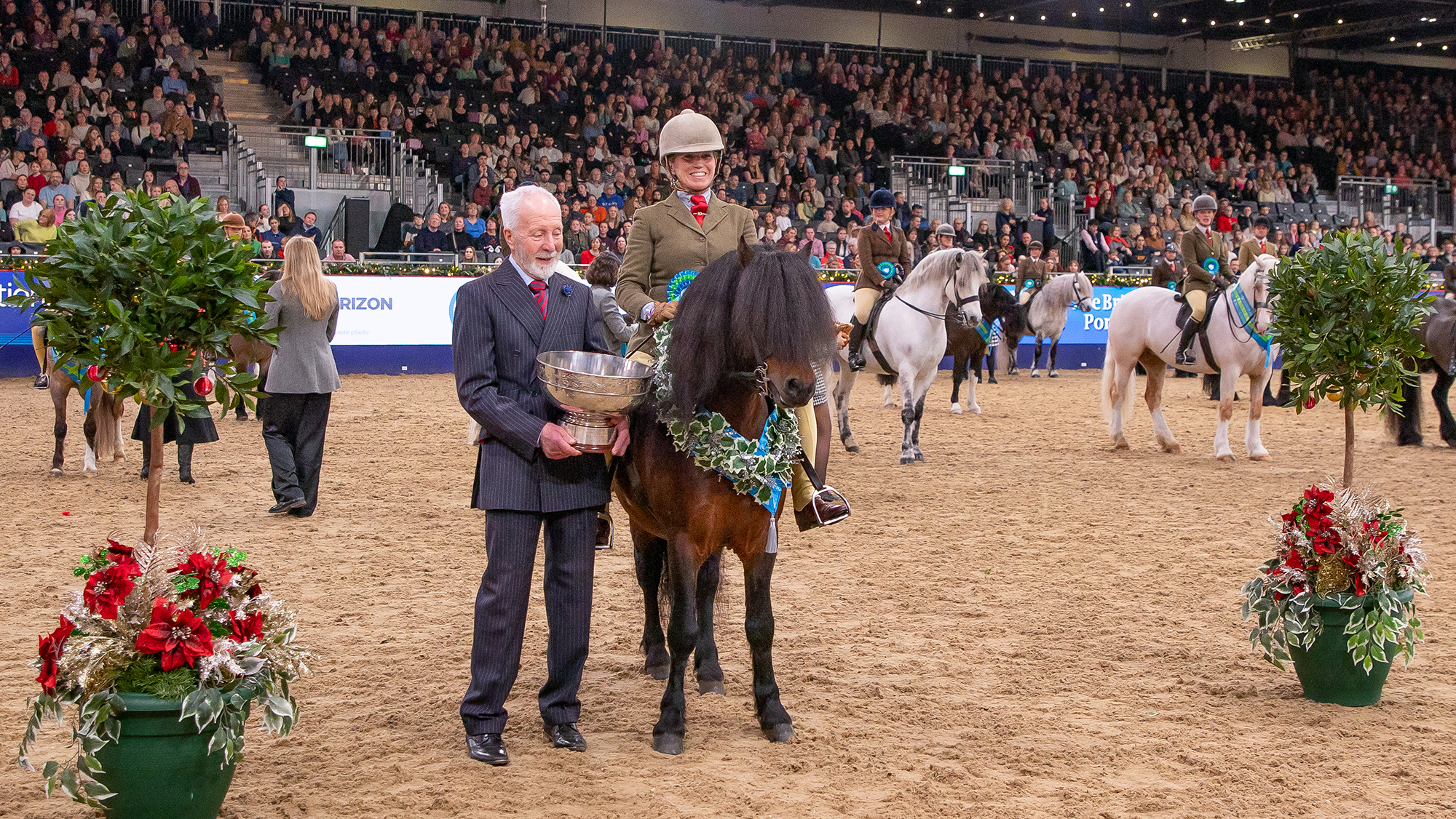 Katie Marriott-Payne smiles at the camera at London International as her M&amp;M ridden champion Salcombe Starehole Bay wears his sash. A man stands alongside with a large punch bowel as the trophy.