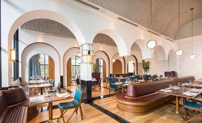 The main room at Origin Grill & Bar, Singapore featuring wood flooring, pendant lights, reddish brown and blue wooden seating and square wooden tables with tableware. The room design features multiple arches