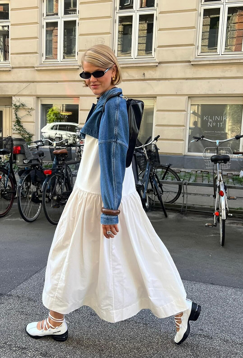 The best ballet sneakers are shown in a photo of a woman standing in the street in Copenhagen wearing a denim cropped jacket over a white maxi dress styled with white lace-up ballerina sneakers