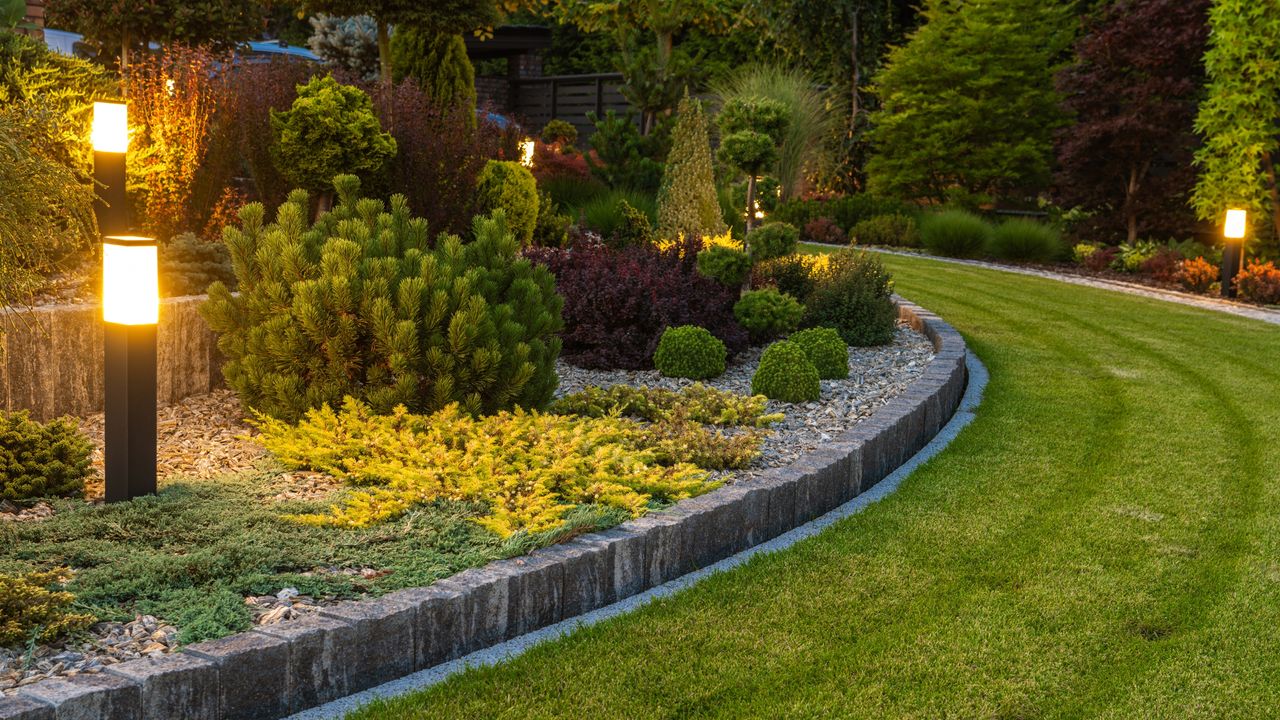 Professionally Landscaped Backyard Garden with Evenly Mowed Lawn and Trimmed Shrubs Illuminated with Outdoor Bollard Lamps - for article on &#039;how much does landscaping cost for small, medium and big yards