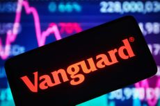 The Vanguard Group logo on a smartphone with a stock chart and ticker board blurred in the background.