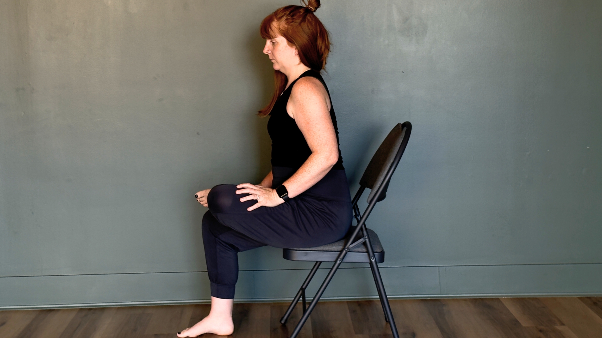 woman performing a seated pigeon pose on a chair on a wooden floor against a sage green wall
