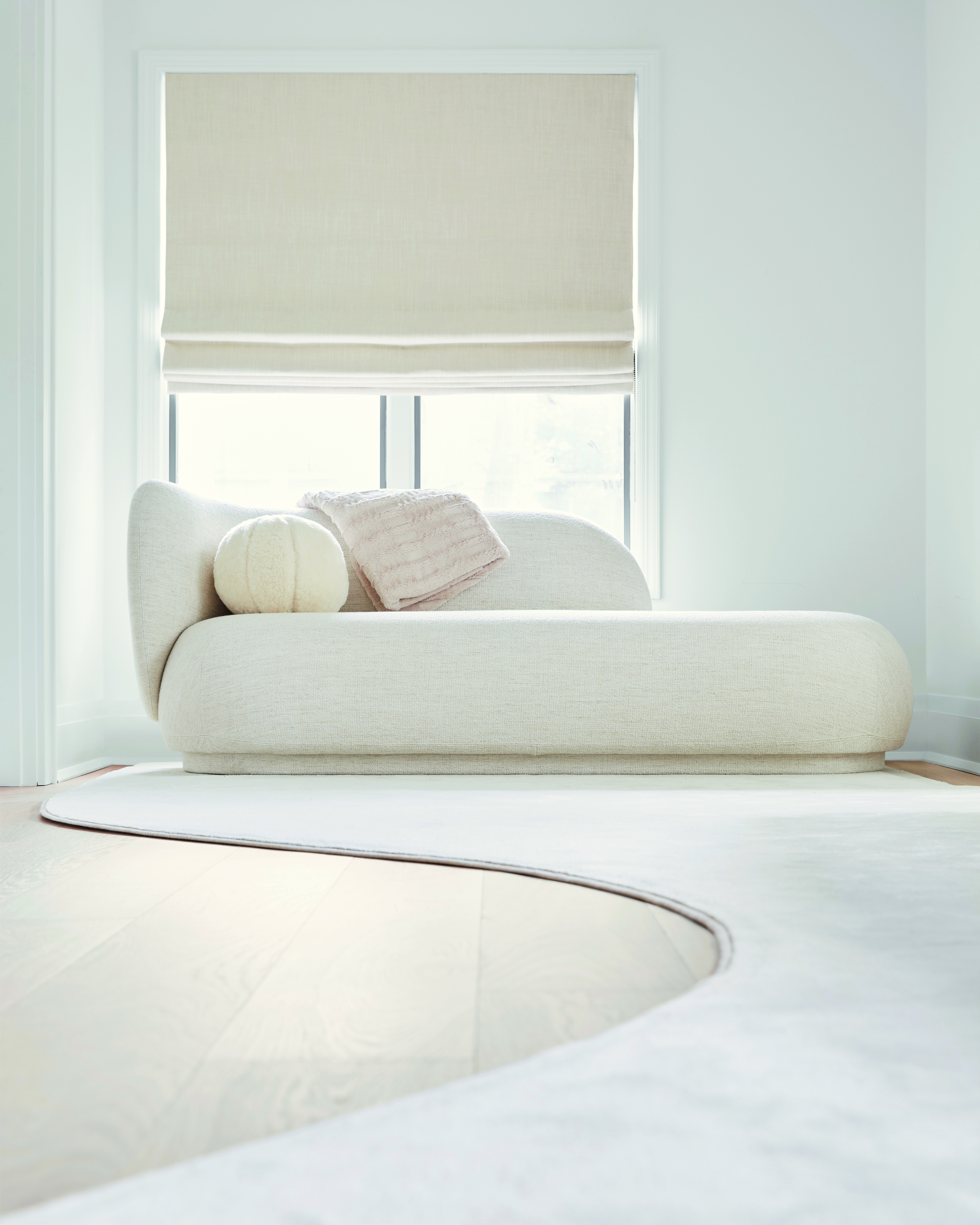 a cream daybed in an all-cream room with pale wood flooring and wavy cream rug and a crem roman blind halfway across the window