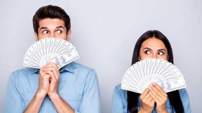 A man and a women hold fans of money in front of their mouths and give each other the side-eye.