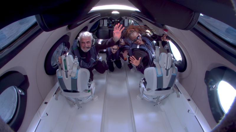 Space tourists wave to the camera inside a spacecraft.